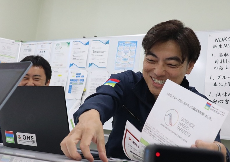 資料を手に笑顔で話す日本ダストの社員。奥でもう一人の社員も笑っている